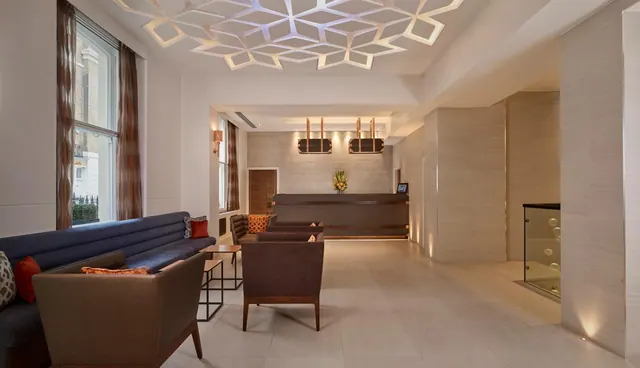 A hotel lobby with upholstered sofas and armchairs, a reception desk in the background, and a geometric lattice ceiling light.