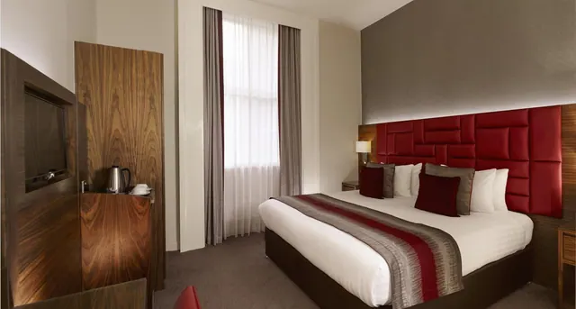 A double bed with a red padded headboard sits beside a wall-mounted television and a wooden cabinet holding a kettle and cups in a hotel bedroom.