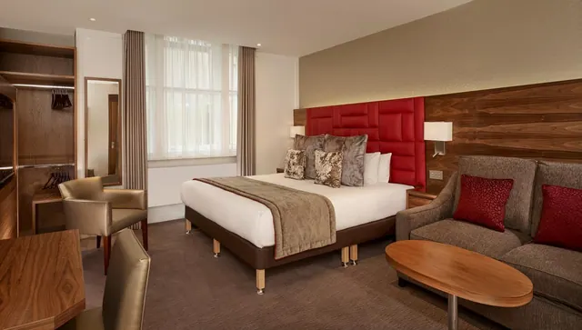 A hotel bedroom with a king bed under a red quilted headboard, a brown sofa with red cushions, a wooden wardrobe and an oval wooden coffee table.
