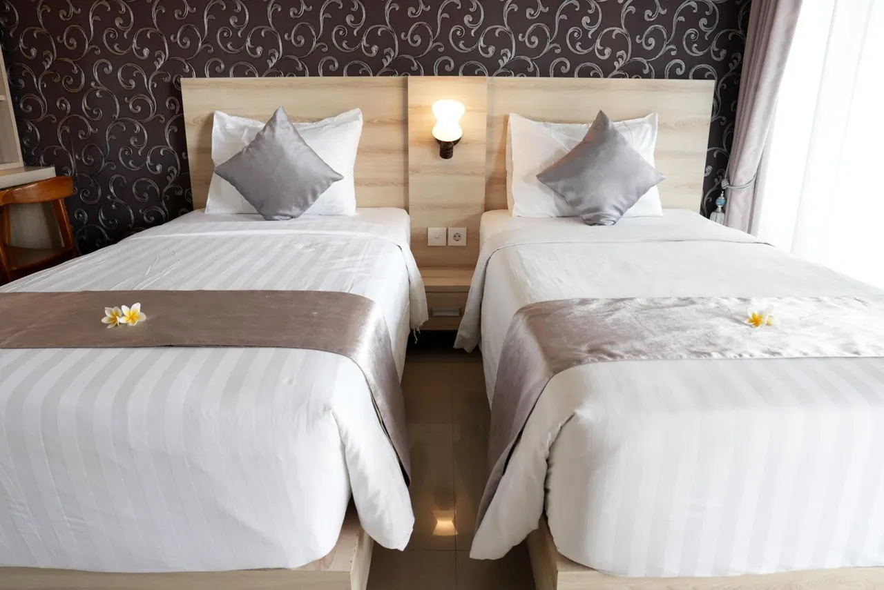 Two single beds with light wooden headboards, white striped linens and grey cushions sit side by side in a bedroom with patterned wallpaper.