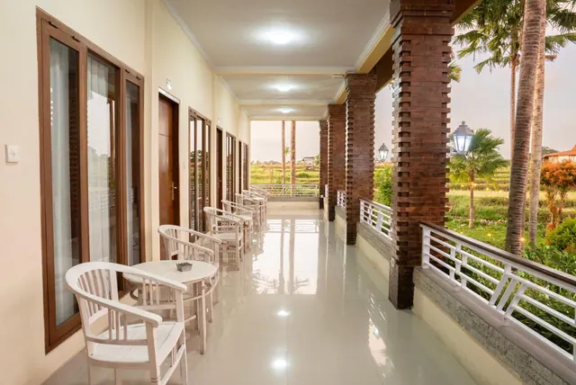 A covered veranda with white chairs and small round tables runs alongside glass doors, with brick pillars and a white railing, and palm trees and rice fields visible beyond.