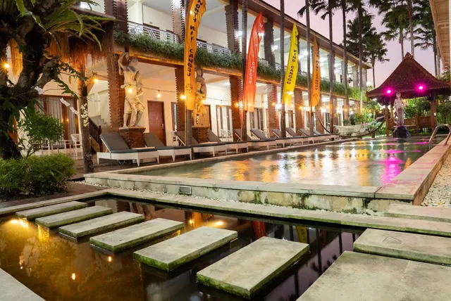 A long pool at dusk with sun loungers along one side, a stepping-stone walkway over a water channel, and a two-storey building with carved wooden columns and hanging flags.