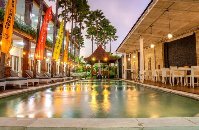 A long swimming pool at dusk with white sun loungers on the left, colourful flags, a gazebo and palm trees beyond, and covered outdoor dining tables on the right.