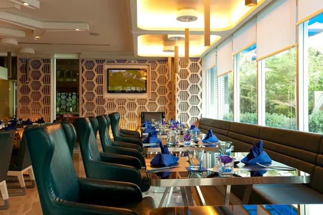 A restaurant dining room with high-back leather chairs, a banquette along large windows looking onto trees, set tables with blue napkins, and a wall-mounted television.