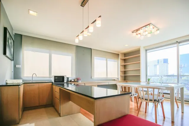 A bright kitchen with an island counter, open shelving, and a dining table with chairs, with tall windows showing city buildings.