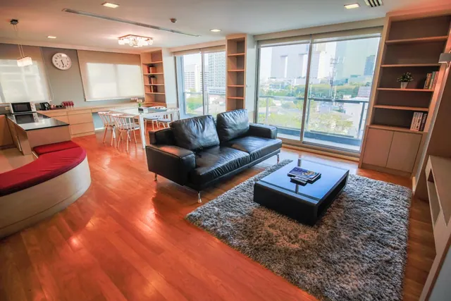A living area with a black leather sofa, a coffee table on a grey rug, a dining table, a kitchen counter, and floor-to-ceiling windows opening onto a balcony with city buildings outside.