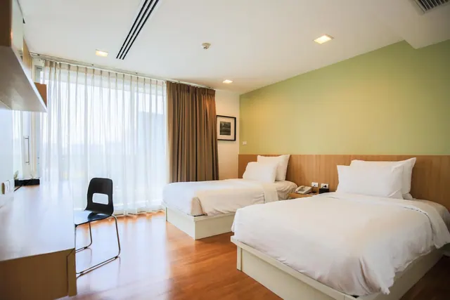 A hotel bedroom with two single beds made with white bedding, a wooden work desk and chair, and a large window with sheer curtains.