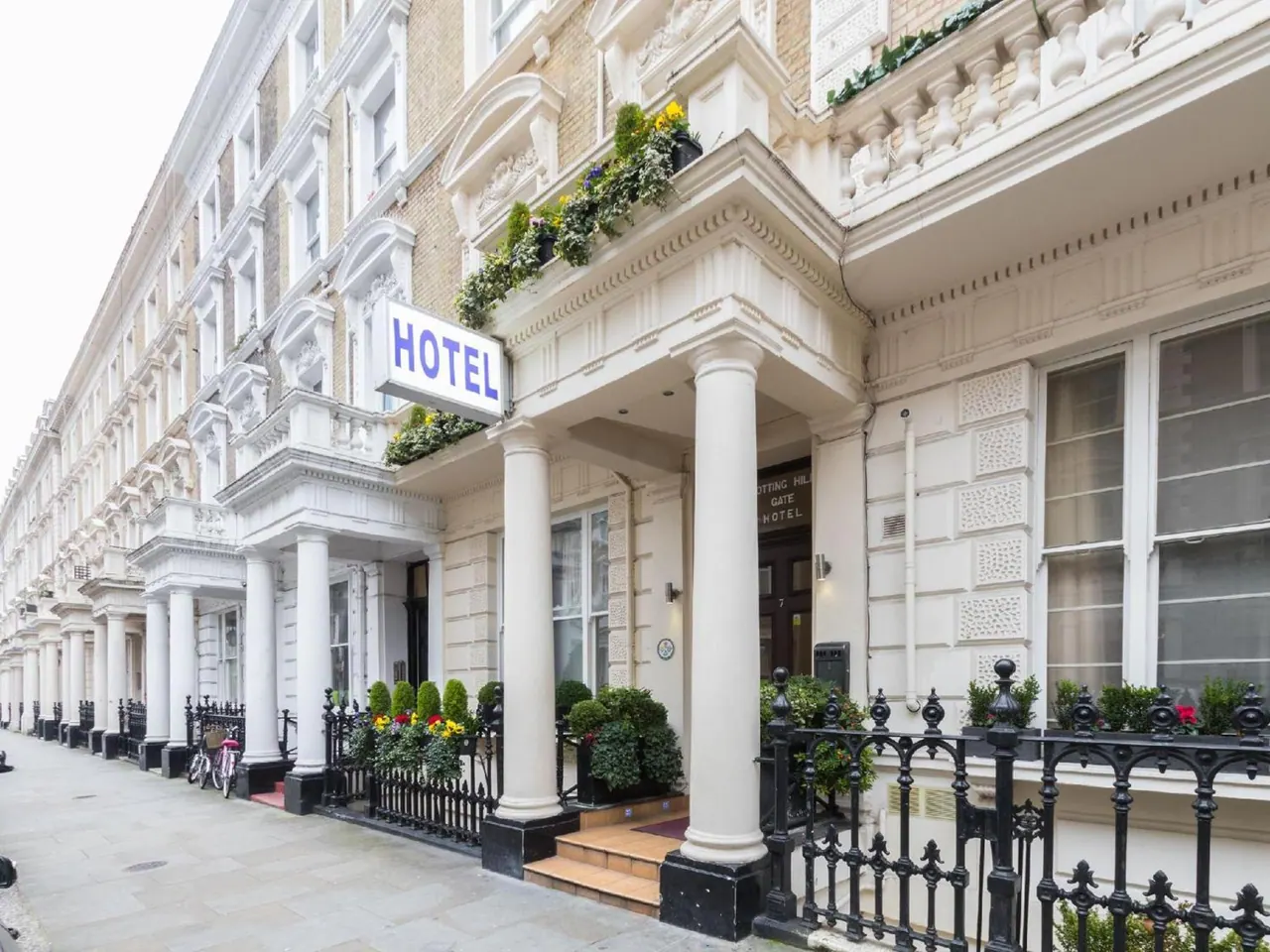 The white stucco facade of a hotel with columned entrance porch, a blue hotel sign, and black iron railings along the pavement.