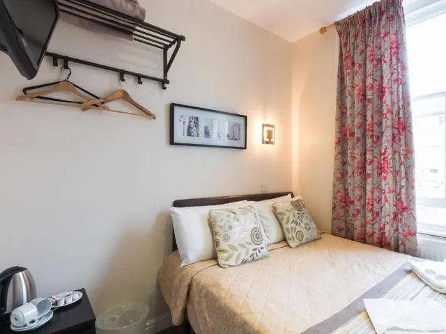 A small hotel bedroom with a double bed, patterned cushions, red floral curtains by the window, a wall-mounted television, and a kettle on a side table.