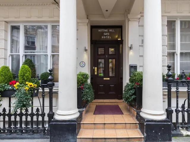 The front entrance of the Notting Hill Gate Hotel, with white columns, a dark wooden door, a red doormat on tiled steps, and potted plants beside wrought iron railings.