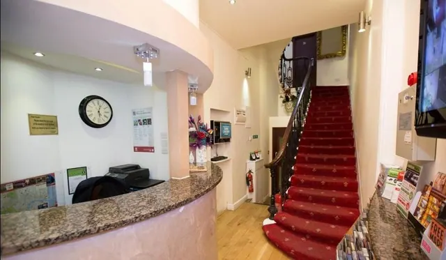 A hotel reception area with a curved granite desk, a wall clock, a printer and a staircase with red carpet.