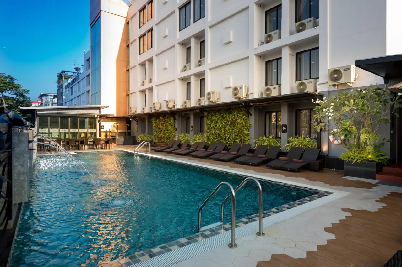 A swimming pool with metal ladders and dark sun loungers along its edge, beside a white multi-storey hotel building with wall-mounted air conditioning units.