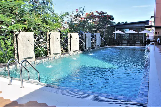 An outdoor swimming pool with water jets spilling from concrete wall panels, with trees behind and a dining area under white umbrellas beyond the pool.