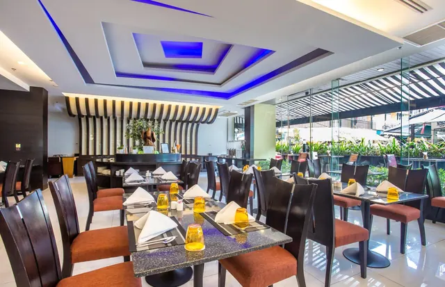 A restaurant dining room with set tables, orange-cushioned dark wood chairs, blue-lit ceiling coffers, and a glass wall looking onto plants outside.
