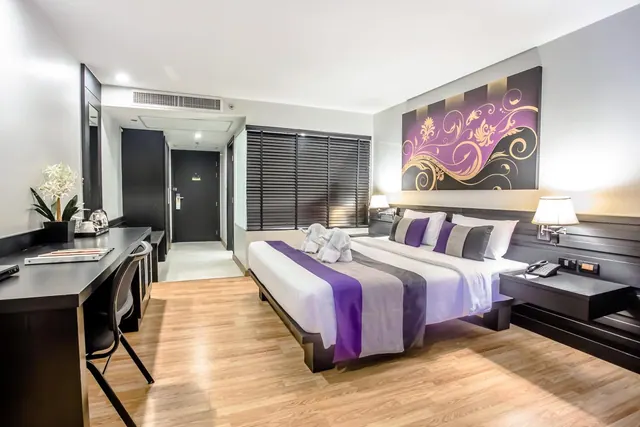 A hotel bedroom with a king bed dressed in purple and grey runners, a work desk with a kettle on the left, and a large decorative wall panel behind the bed.