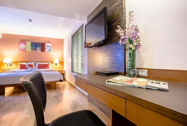 A hotel bedroom with a double bed, a wall-mounted television above a wooden desk holding brochures and a vase of flowers, and an office chair in the foreground.