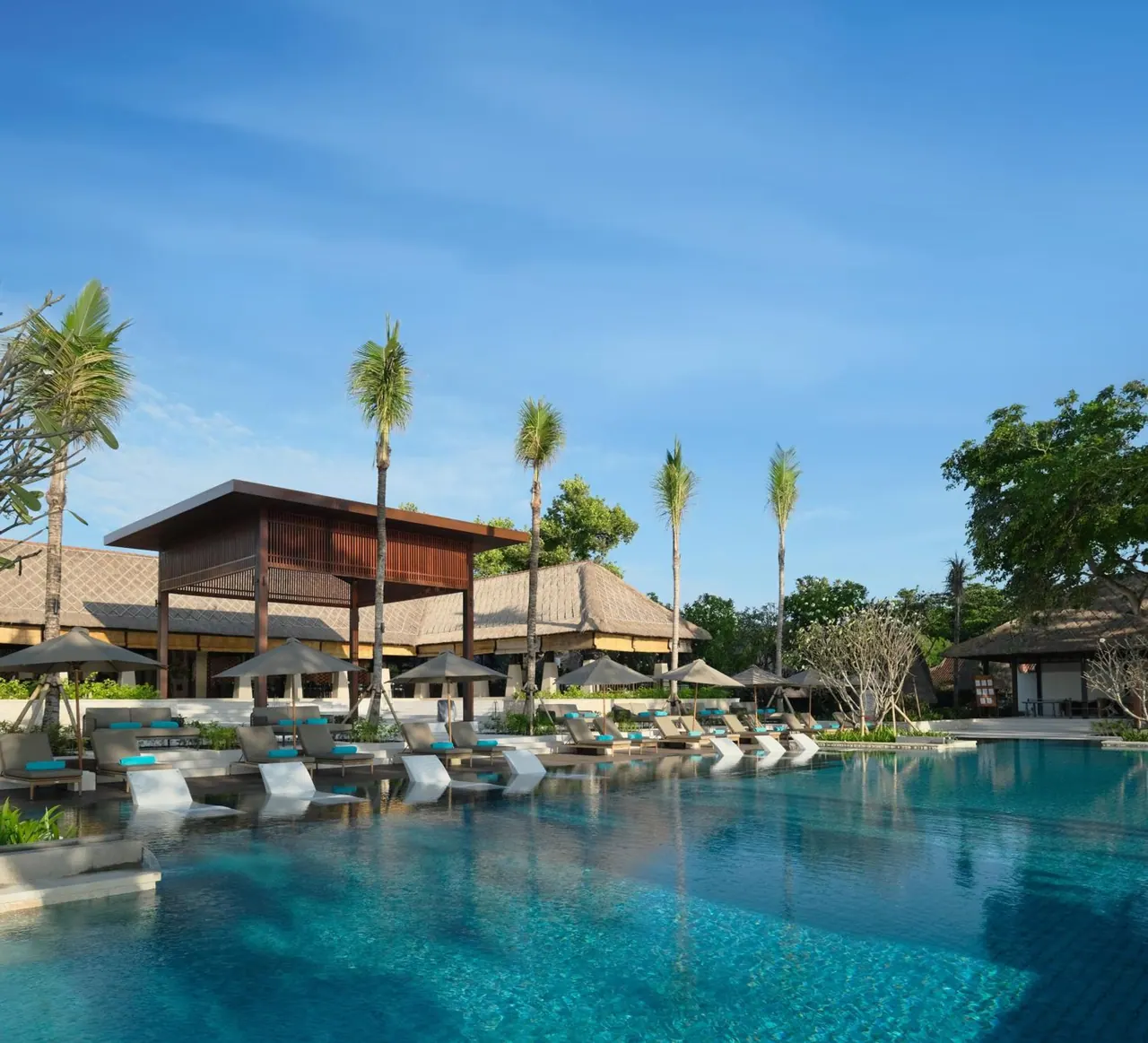 A large swimming pool with rows of sun loungers and grey parasol umbrellas, with palm trees and thatched-roof pavilions beside it under a clear blue sky.