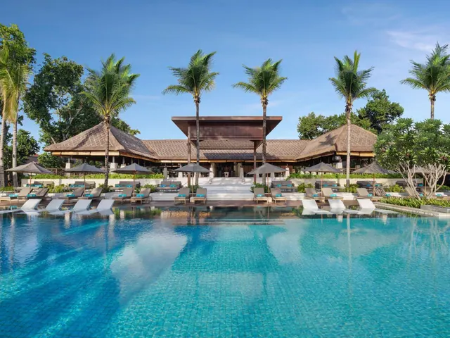 A large turquoise swimming pool sits in front of a thatched-roof pavilion, with sun loungers, umbrellas and palm trees around it.