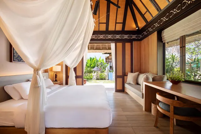 A bedroom with a made bed under sheer canopy drapes, a cushioned bench, a wooden desk and chair, and a window looking out onto tropical garden plants.