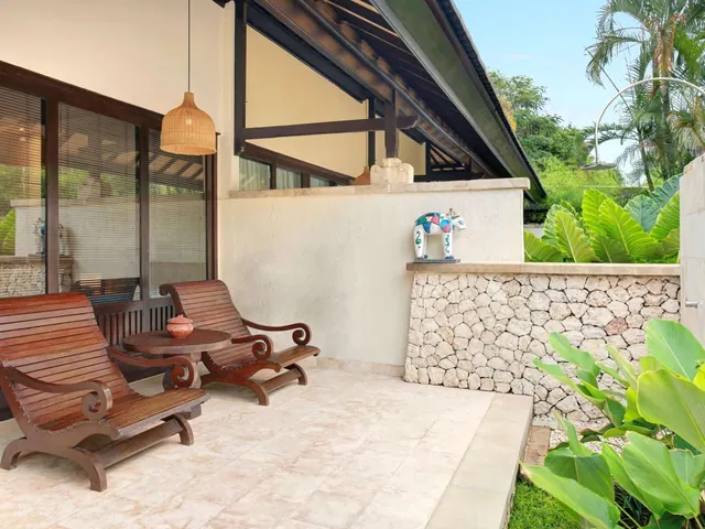 A covered terrace with two wooden lounge chairs, a round wooden table and a rattan pendant lamp beside a stone wall and tropical plants.
