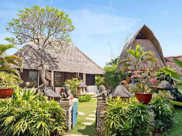 A stone-paved garden path lined with tropical plants and stone statues leads toward thatched-roof bungalows under a clear blue sky.