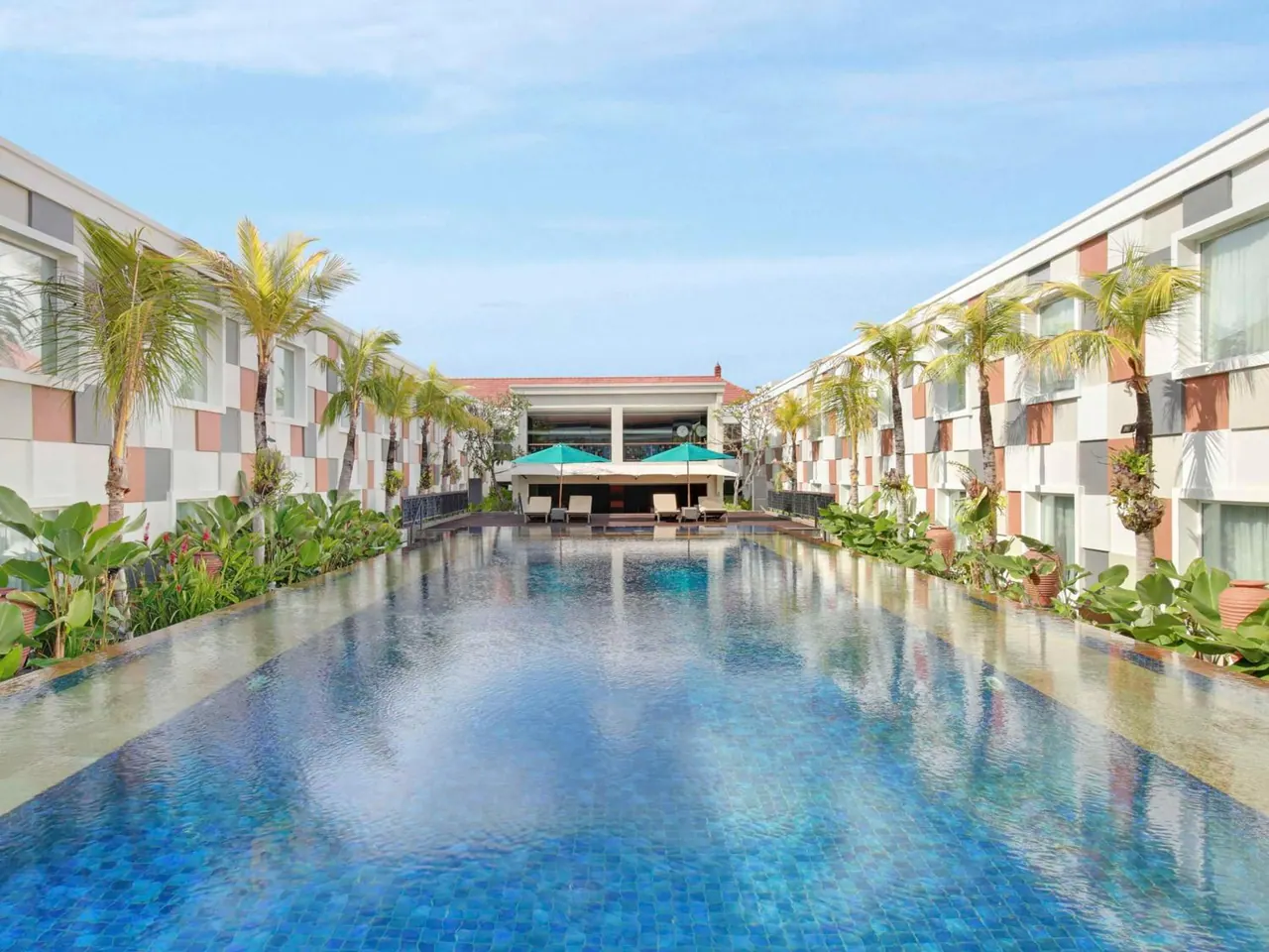 A long blue-tiled lap pool runs between two white buildings lined with palm trees, with teal umbrellas and loungers at the far end.