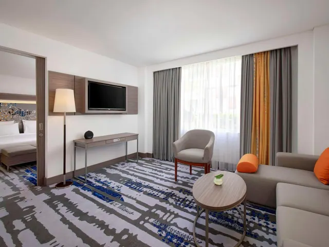 A hotel suite living area with a sofa, armchair and wall-mounted television, with a bed visible through the open doorway on the left.