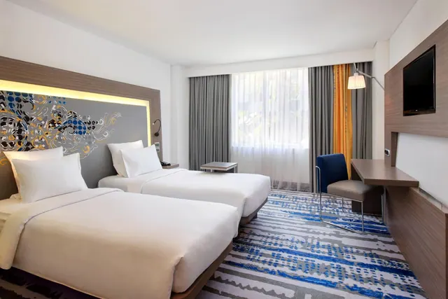 A twin-bedded hotel room with a patterned headboard, a window with sheer and grey curtains, a desk, a blue chair and a wall-mounted television.