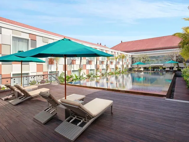A wooden deck beside a long swimming pool holds wicker sun loungers and teal parasols, with a white hotel building behind.