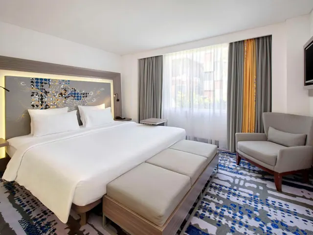 A hotel bedroom with a large king bed, a bench at the foot of the bed, an armchair by a window with sheer curtains, and a wall-mounted television.