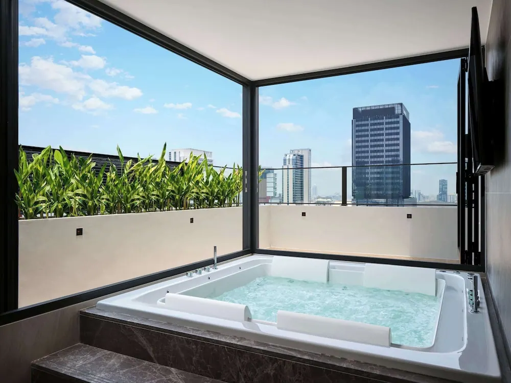A built-in jacuzzi sits on a private terrace with floor-to-ceiling glass, looking out over a city skyline with tall office towers under a blue sky.