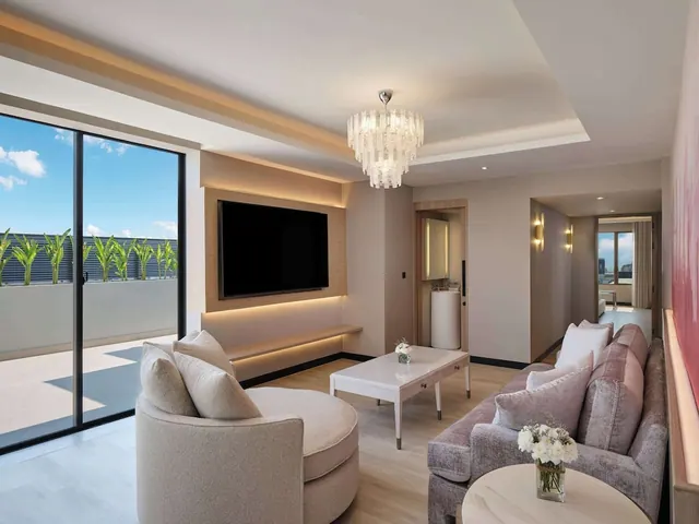 A living room with a wall-mounted television, a curved armchair, a sofa, a chandelier, and a glass sliding door opening onto a balcony with potted plants.