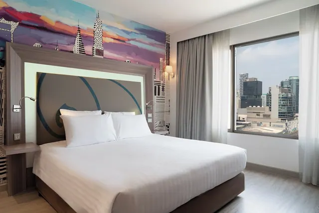 A bedroom with a made king bed, a painted wall mural of a city skyline at sunset, and a window looking out over high-rise buildings.