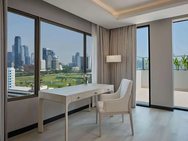 A bright room with a white desk and armchair beside large windows overlooking a city skyline and a green golf course, with a balcony on the right.