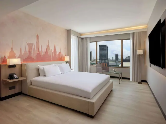 A made king bed with white linen sits in a room with a painted temple mural on the wall, a wall-mounted television, and a large window overlooking city buildings.
