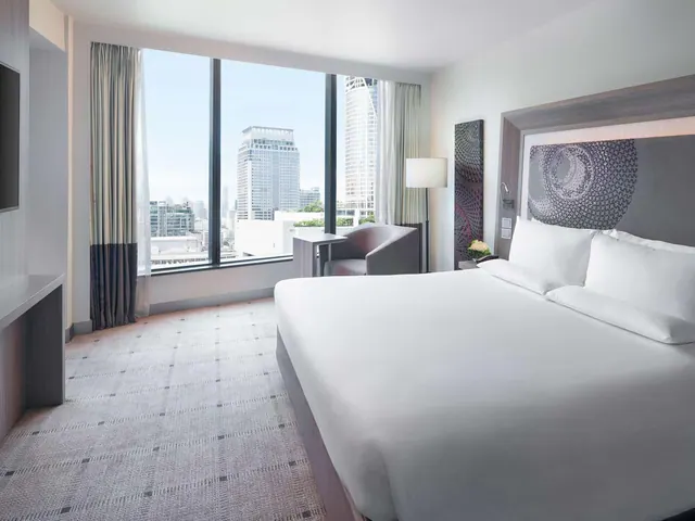 A bedroom with a large white king bed facing floor-to-ceiling windows that look out over city high-rise buildings.