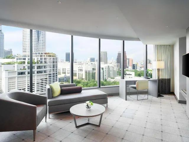 A hotel suite living area with a grey sofa, armchair, round coffee table, writing desk and wall-mounted television, with floor-to-ceiling windows looking over city high-rises and trees.