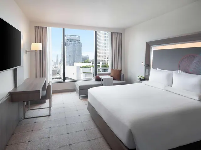A king bed, work desk and seating area sit in a hotel bedroom facing a large window with city towers outside.