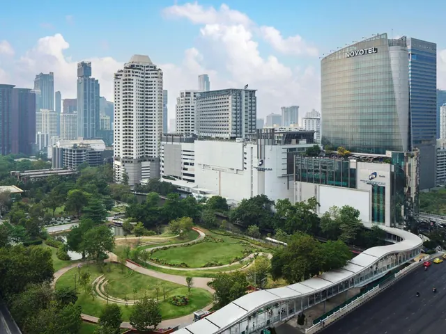Aerial view of a curved glass hotel tower with a Novotel sign, beside a landscaped park and a covered elevated walkway, with city towers behind.