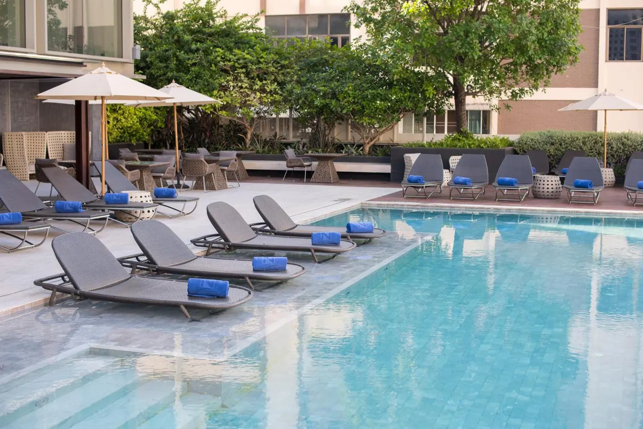 A large pool with grey sun loungers holding rolled blue towels, shaded by white parasols, with trees and a hotel building behind.