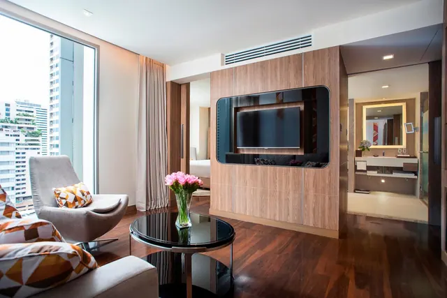 A hotel suite living area with a wall-mounted television, an armchair, a round glass coffee table holding pink flowers, and a tall window onto city buildings, with doorways leading to a bedroom and a bathroom.