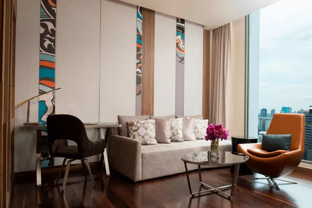A living area with a beige sofa, orange armchair, glass coffee table and desk beside a floor-to-ceiling window overlooking city buildings.