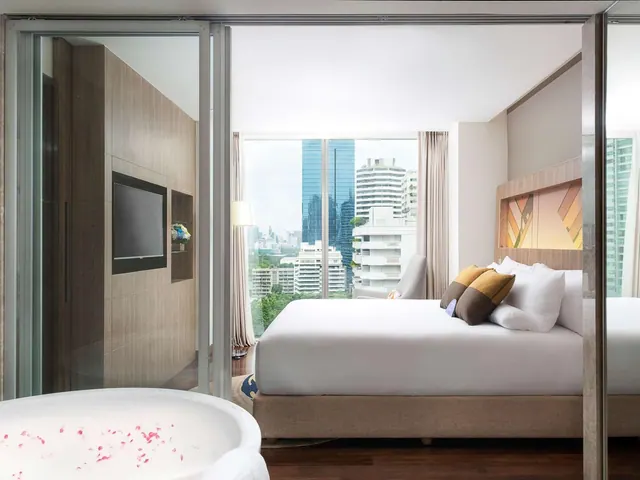 A bedroom with a large bed, a freestanding bathtub with rose petals in the foreground, a wall-mounted television, and a window overlooking city high-rise buildings.