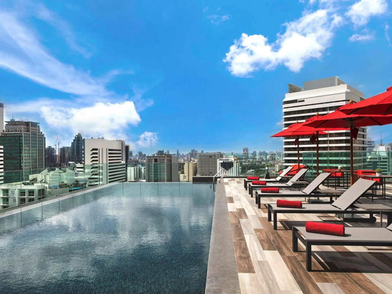 A rooftop infinity pool with grey sun loungers, red cushions and red umbrellas overlooks a city skyline under a blue sky.