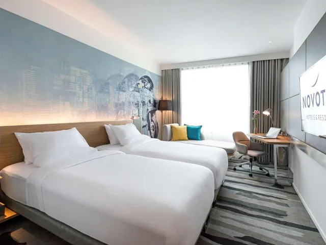 A hotel bedroom with two white beds, a grey sofa with cushions, a desk with a chair and lamp, and a wall-mounted television.