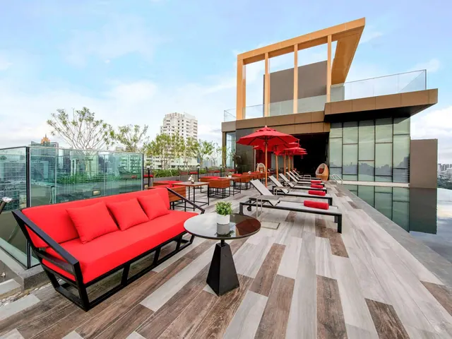 A rooftop terrace with a red sofa, red parasols over sun loungers, and a glass-edged pool, with city buildings and trees visible beyond the railing.