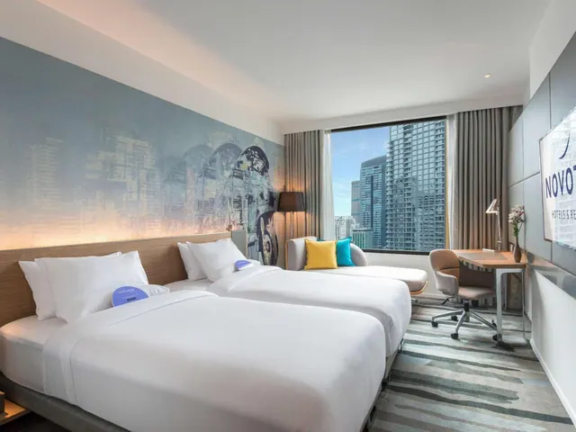 A hotel bedroom with two beds, a seating area with cushions, a desk and chair, and a window showing high-rise towers under a blue sky.