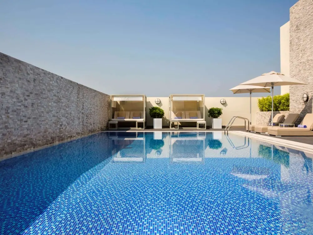 A rooftop swimming pool with blue mosaic tiles, canopied daybeds, sun loungers and parasol umbrellas beside a stone wall under a clear sky.