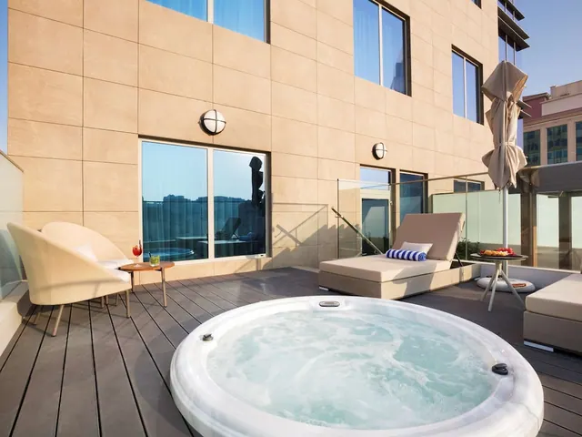 A terrace with a round hot tub, a sun lounger, armchairs and a closed parasol beside a tiled building wall, with buildings visible at the right edge.