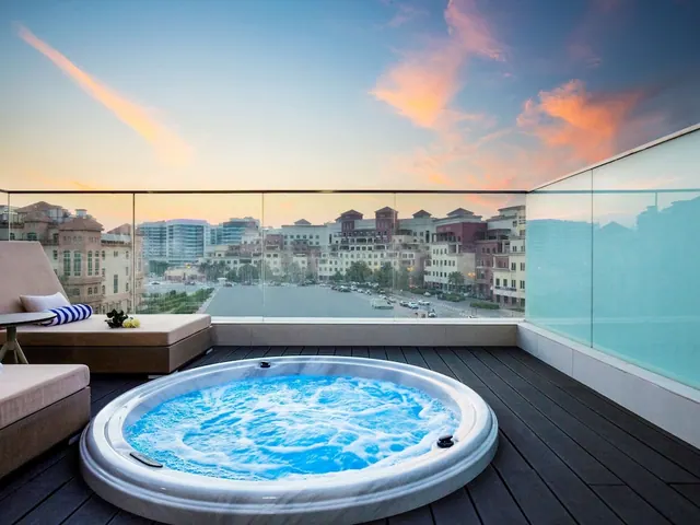 A round hot tub with bubbling blue water sits on a balcony beside loungers, overlooking a city street and low-rise buildings at sunset.